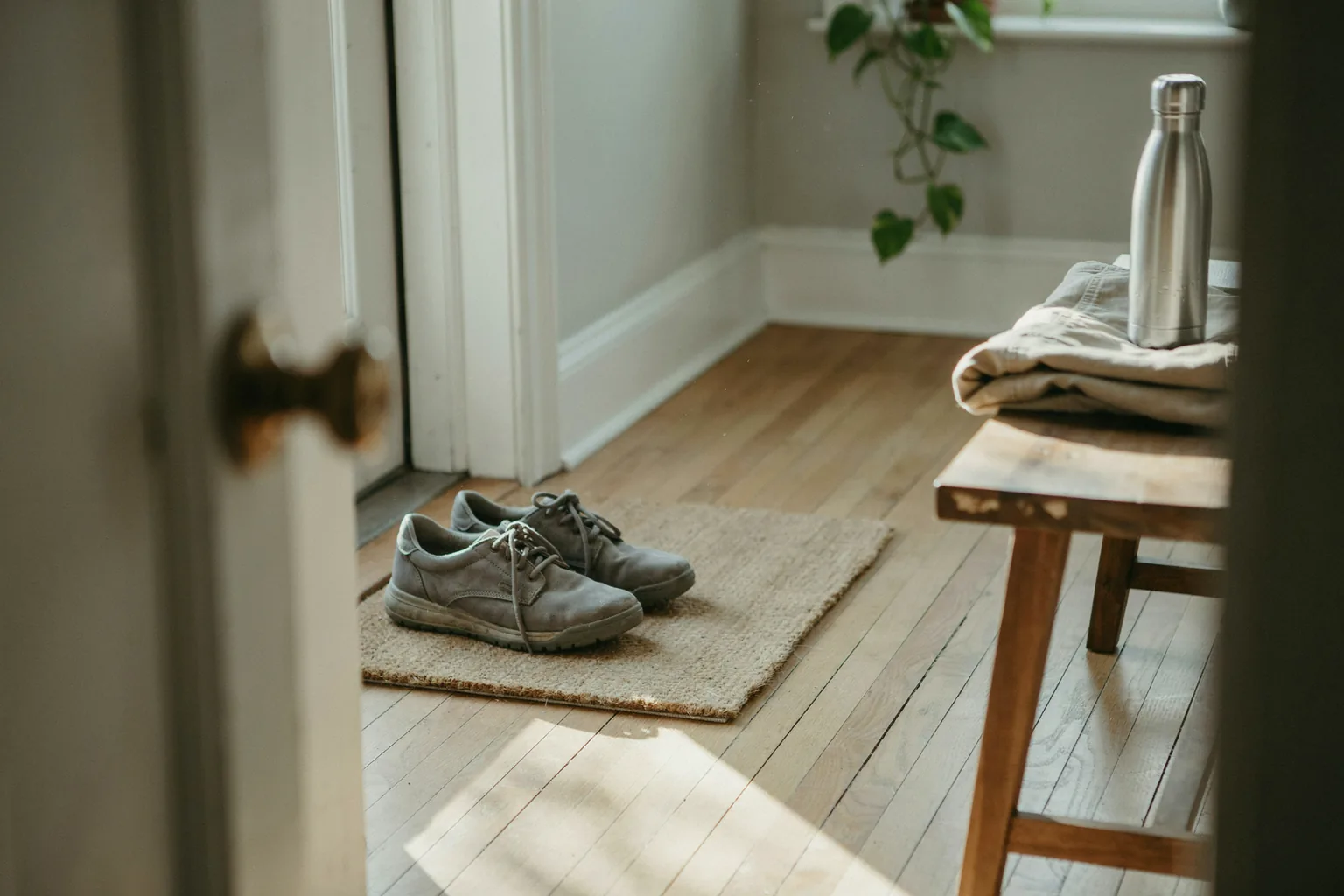 Gray walking shoes on a doormat inside a front door, with a folded jacket and a steel water bottle on a wooden bench
